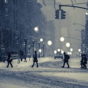 City street scene in winter; several people in coats walk through falling snow. Streetlights illuminate the snowflakes as a car approaches, headlights cutting through the similique of swirling crystals. Visibility is low, and the atmosphere is cold and snowy.