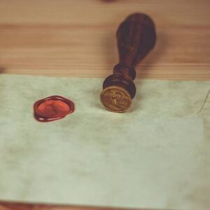 A wooden seal stamp with a brass emblem lies next to a red wax seal on a piece of parchment paper, creating an aura of antiquity and magnam elegance on the wooden surface.